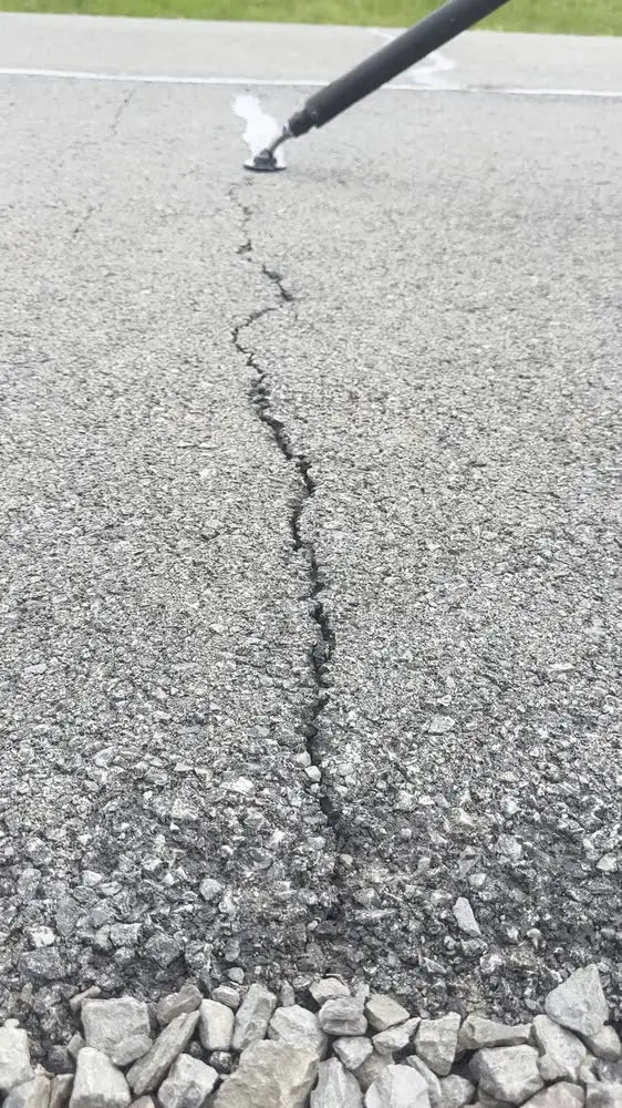 A crack-seal wand running hot rubber sealant into a crack across a gray asphalt surface