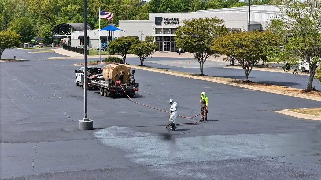 Aerial view of two Arkansas Pavement crew members squeegeeing sealcoat across a church parking lot, the treated half noticeably darker